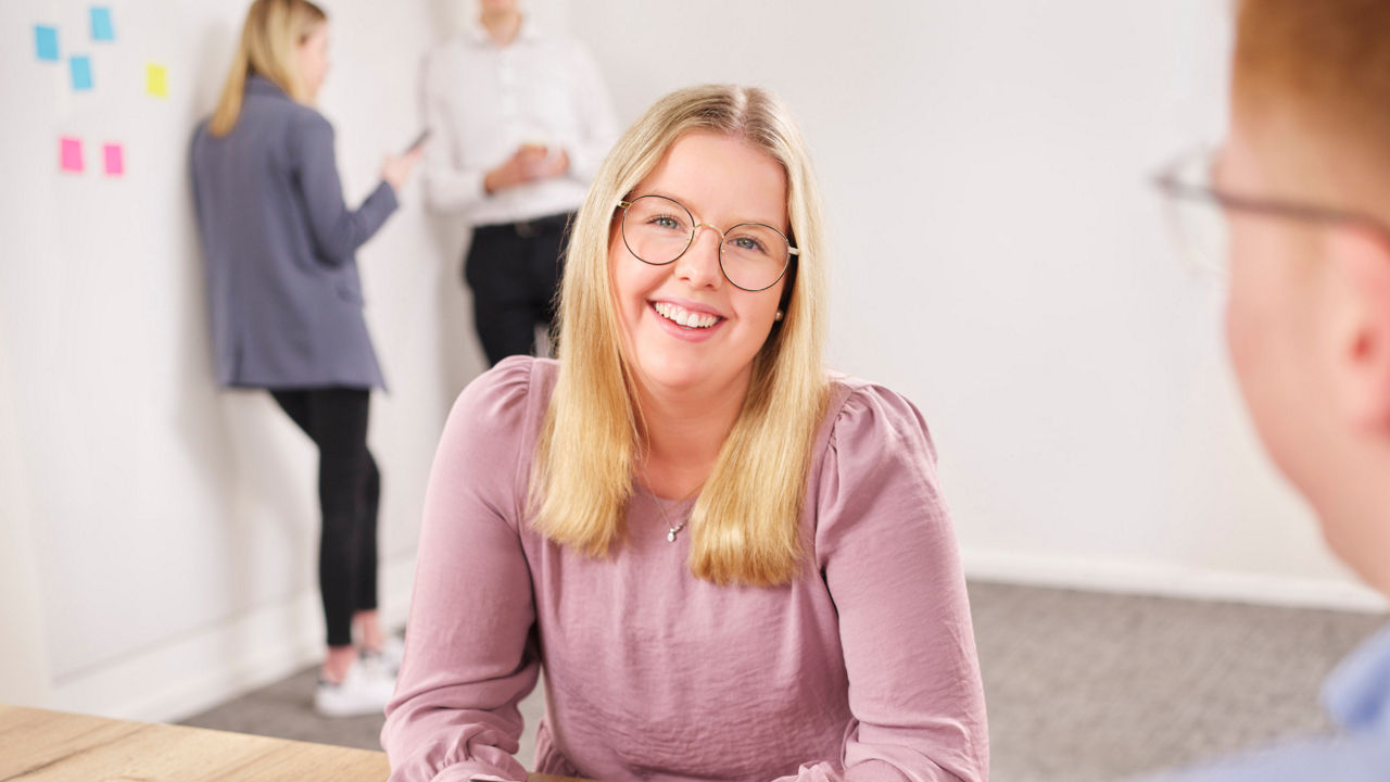 Eine junge Frau sitzt lächelnd am Tisch im Besprechungsraum. Im Hintergrund unterhalten sich zwei Personen.