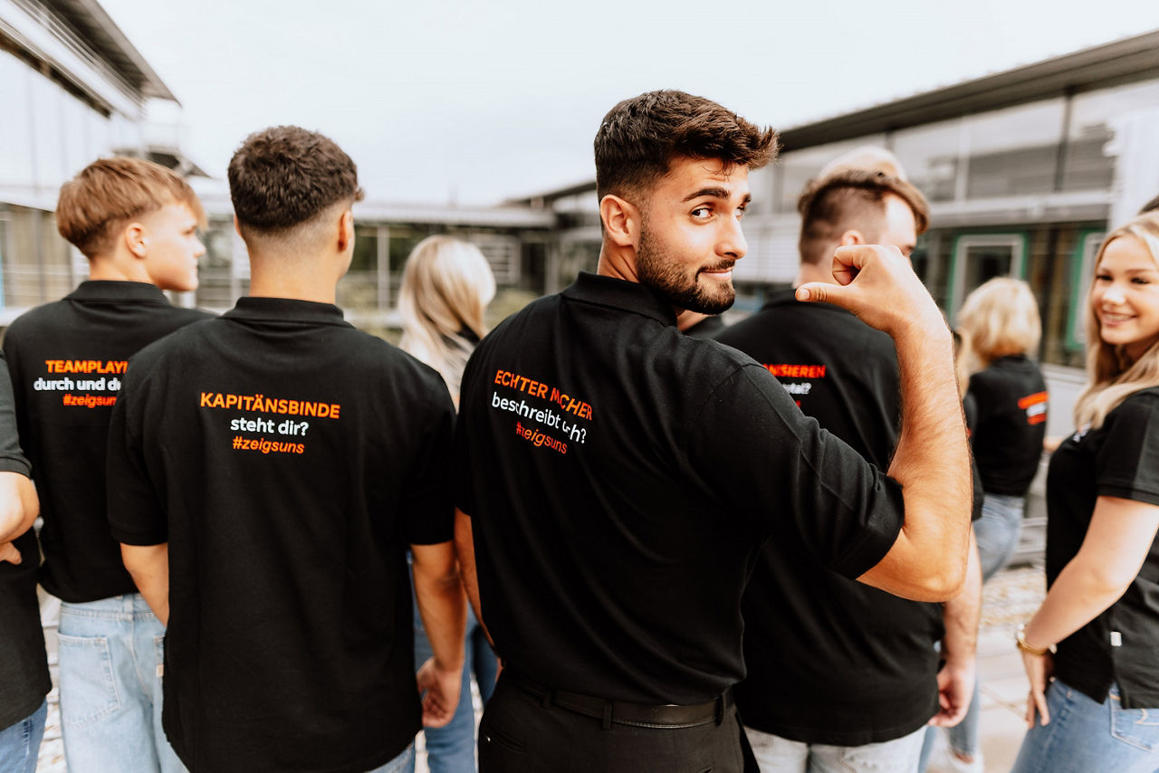 Gruppe junger Menschen mit schwarzen Poloshirts auf deren Rücken in orange ihre Passion steht, zum Beispiel: Teamplayer durch und durch? zeigs uns