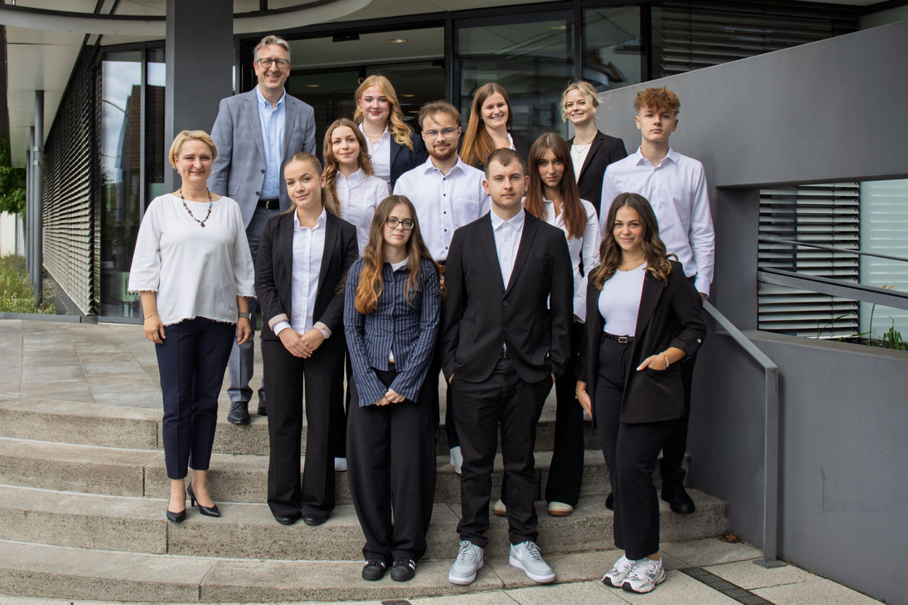 Das Bild zeigt die neuen Auszubildenden, die Bereichsleiterin Personal und den Vorstandssprecher vor dem Bankgebäude. Die Personen stehen in drei Reihen hintereinander auf der Außentreppe. 