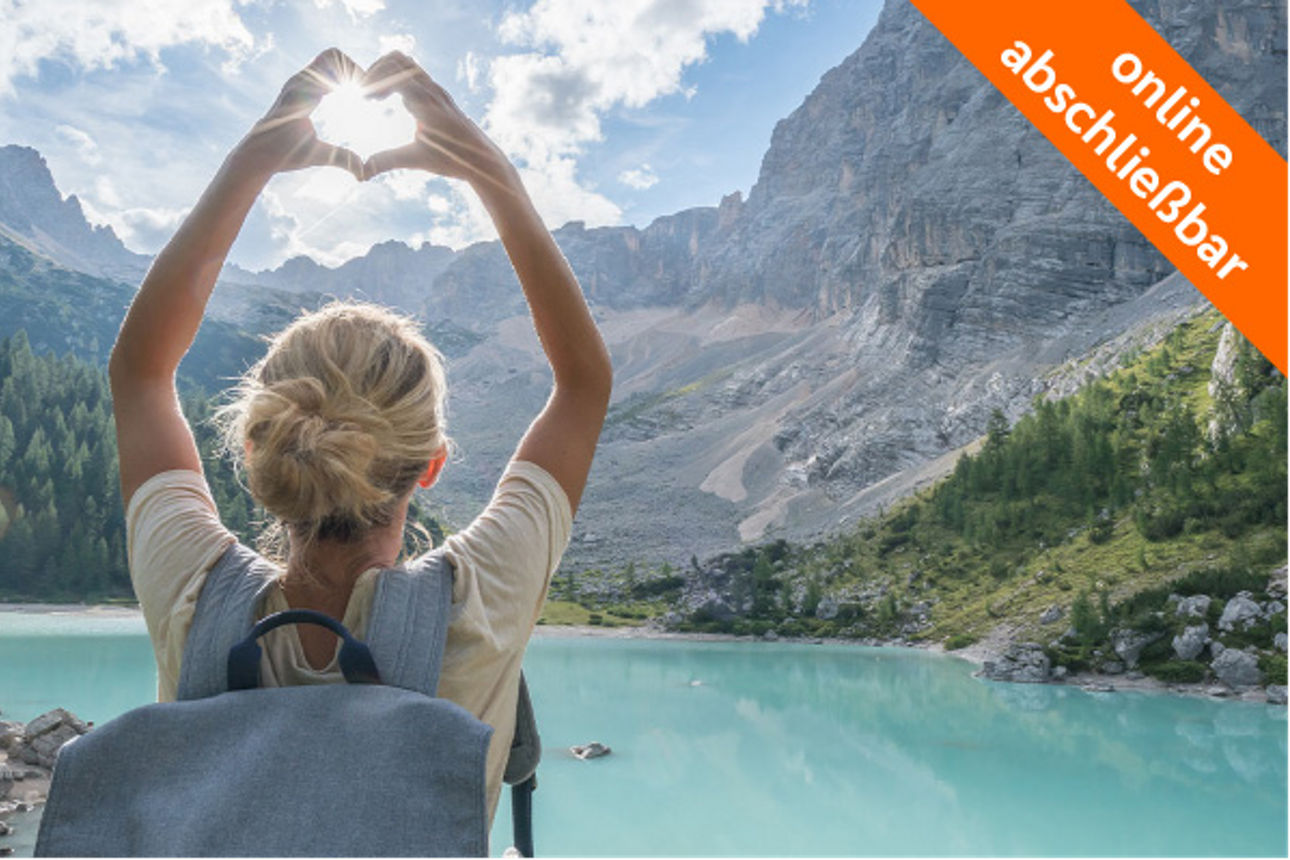 Frau mit Rucksack formt vor einem türkisfarbenen Bergsee ein Herz mit den Händen gegen die Sonne. Oben rechts ein orangefarbenes Banner mit dem Text ‚online abschließbar‘.