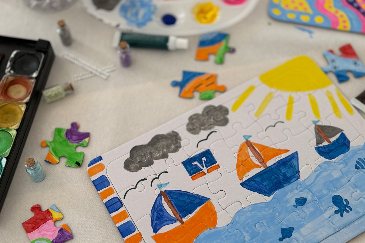 Ein selbst gestaltetes Puzzle mit Segelschiffen, Sonne und Meer, umgeben von Malkasten und bunten Puzzleteilen.