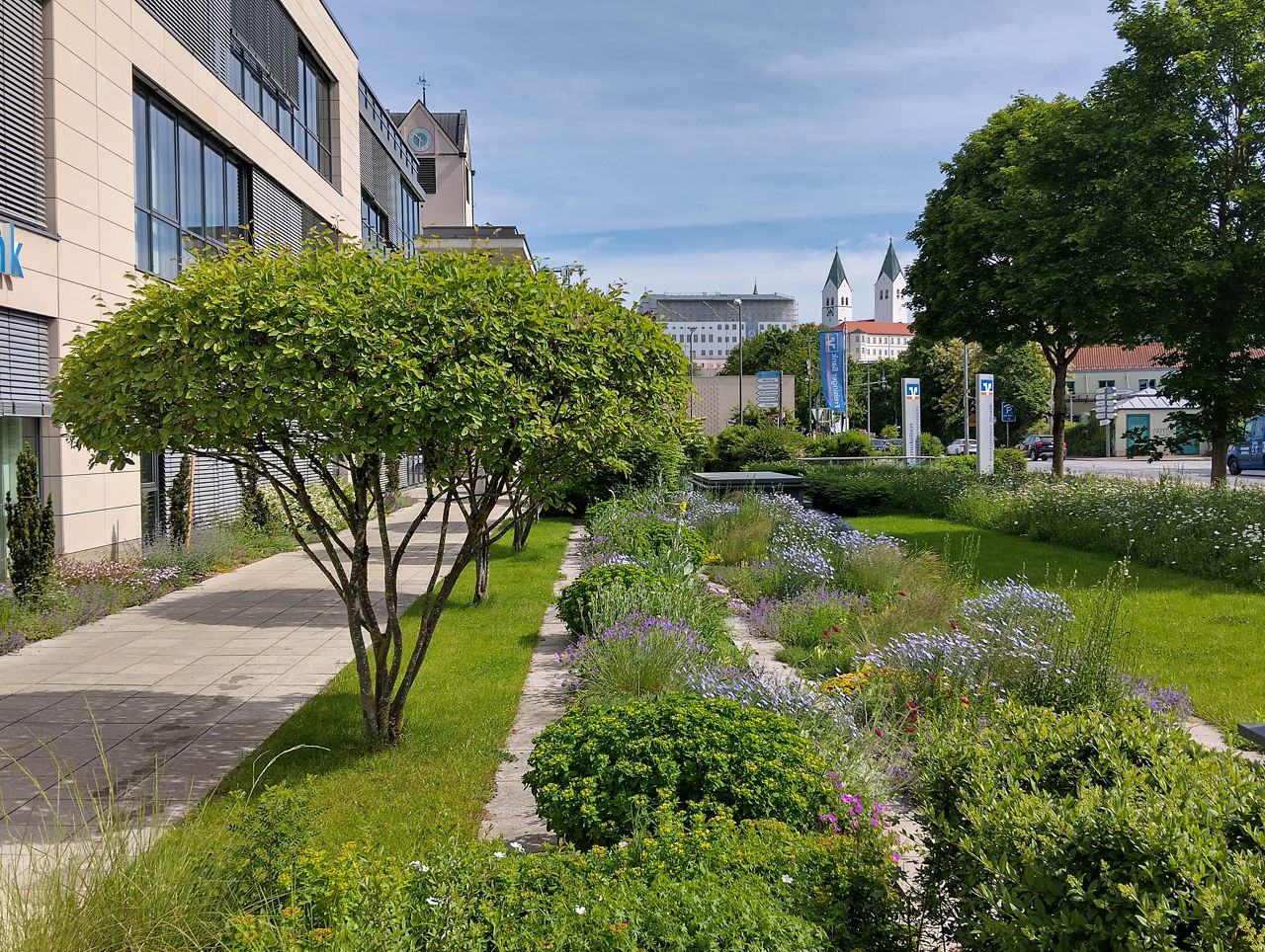 modernes Bankgebäude links neben Gartenanlage mit Felsenbirnbäumen und blühenden Rabatten, im Hintergrund der Freisinger Dom