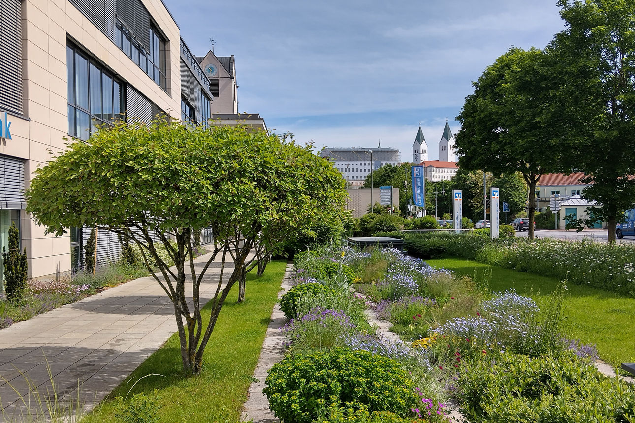 modernes Bankgebäude links neben Gartenanlage mit Felsenbirnbäumen und blühenden Rabatten, im Hintergrund der Freisinger Dom