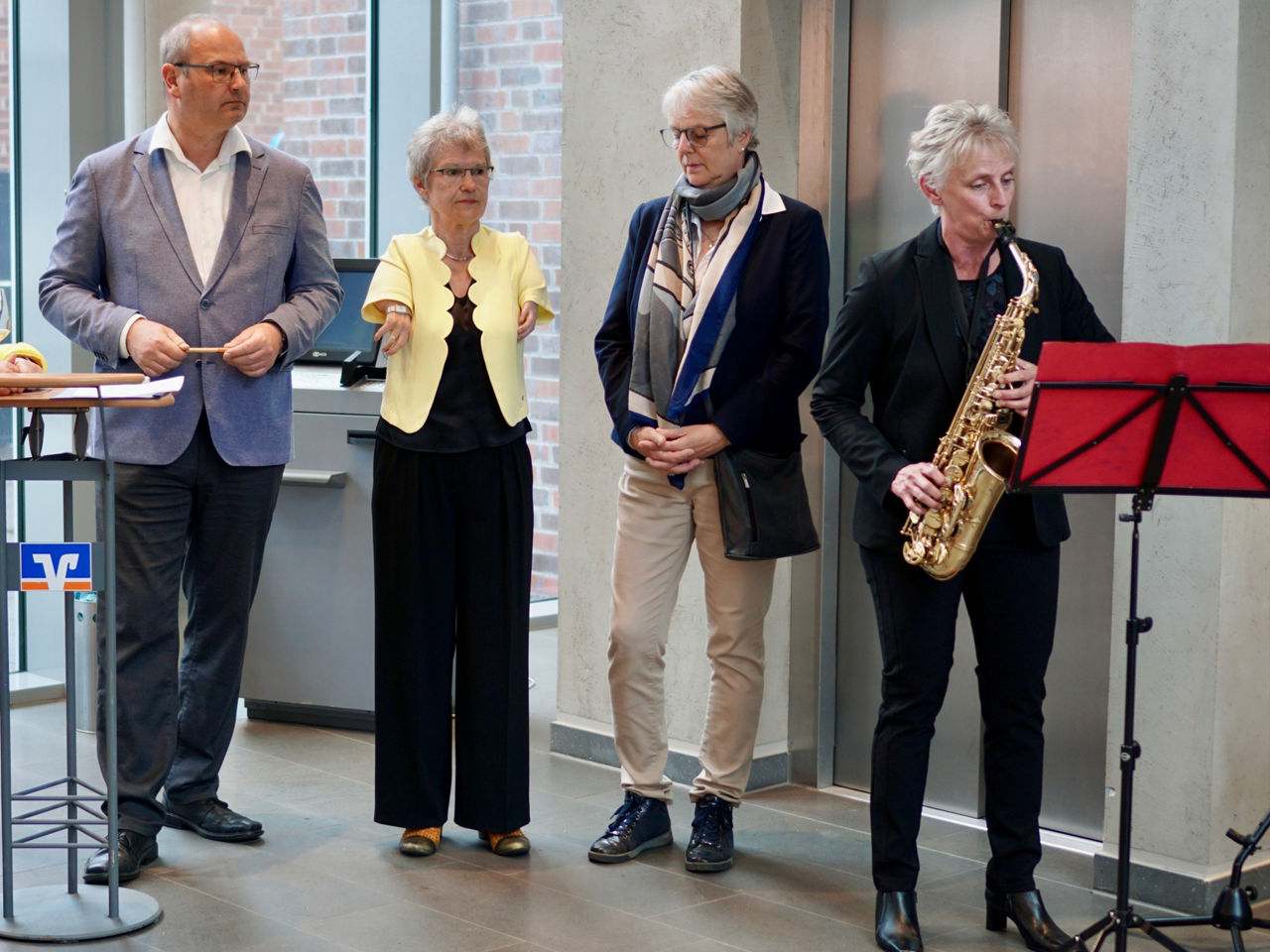 Im Foyer der Volkbank Schermbeck spielt Anja Heix Saxophon. Die Künsterlinnen M. Starke und G. Rott sowie W. Lensing hören aufmerksam zu.