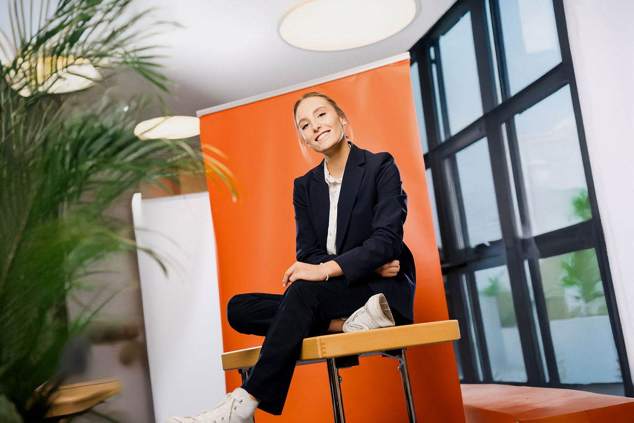 Junge Auszubildende sitzt vor orangefarbenem Rollup und lächelt in die Kamera