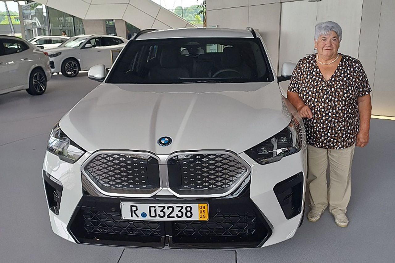 Ältere Dame steht lächelnd neben ihrem Hauptgewinn, einem neuen weißen BMW iX2 in dem modernen Gebäude der BMW-Welt München