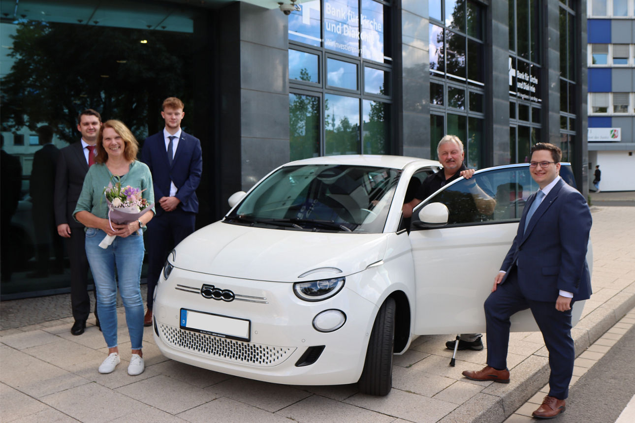 Gewinner-Ehepaar steht vor einem weißen Auto und freut sich über den Gewinn