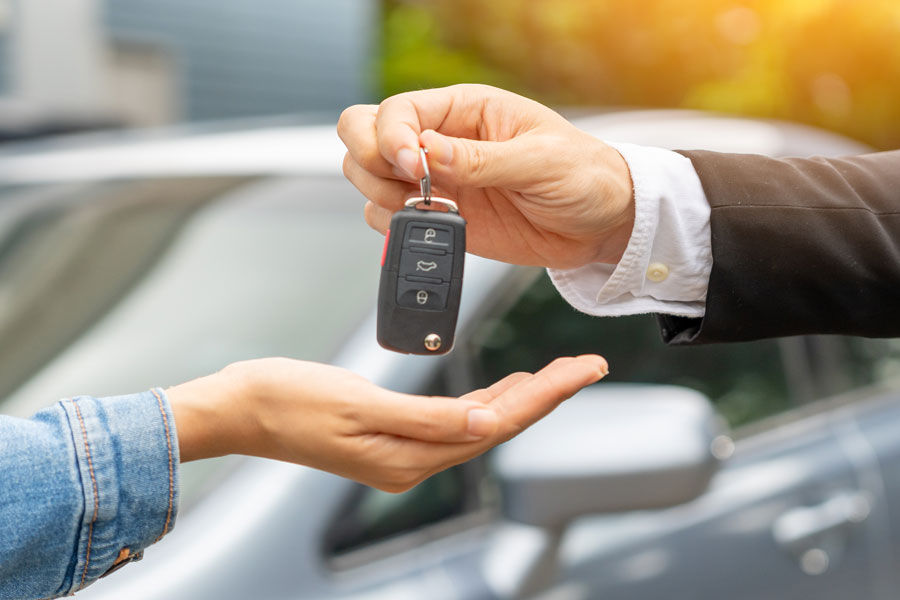 Eine männliche Hand reicht einer weiblichen Hand einen Autschlüssel, im Hintergrund ist ein Auto