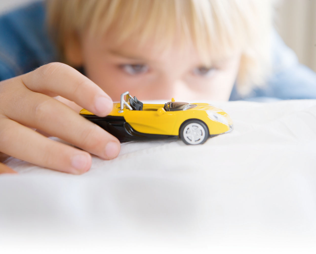 Blonder Junge im Hintergrund (unscharf), der im Vordergrund mit einem gelben Miniaturauto spielt