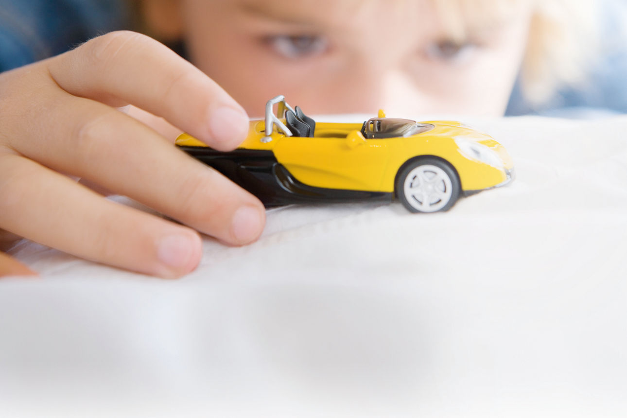 Blonder Junge im Hintergrund (unscharf), der im Vordergrund mit einem gelben Miniaturauto spielt