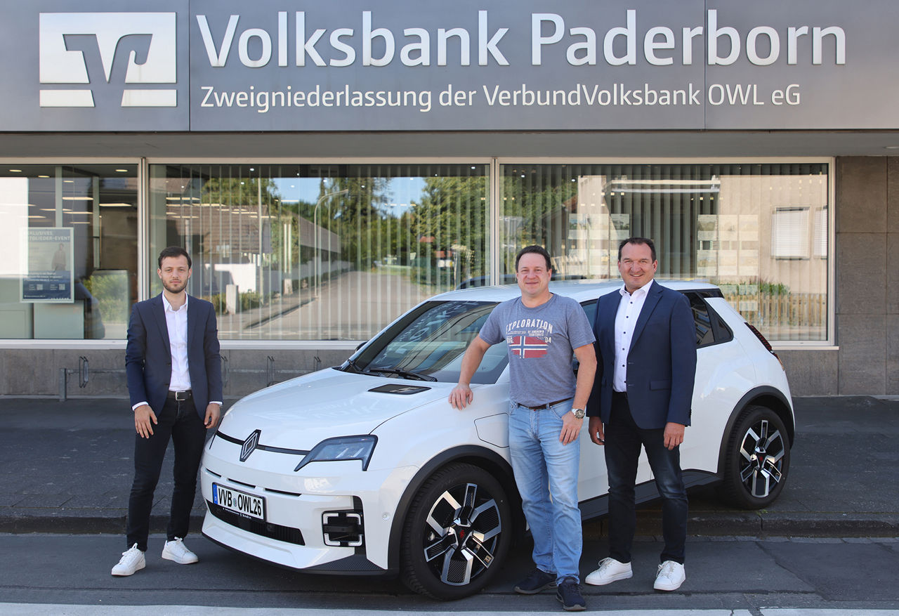 Drei Herren stehen neben einem weißen Elektroauto vor der Filiale der Volksbank Paderborn. gewonnen hat.  