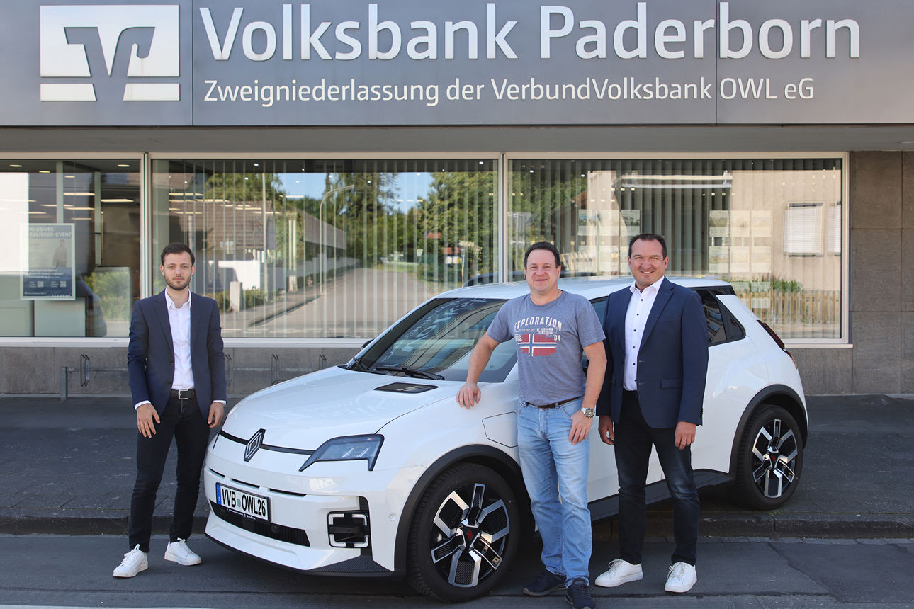 Drei Herren stehen neben einem weißen Elektroauto vor der Filiale der Volksbank Paderborn. gewonnen hat.  