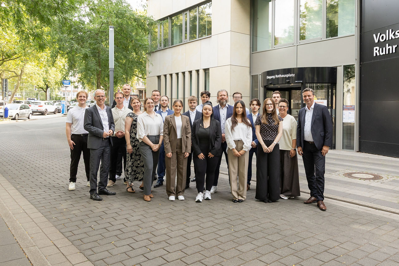 Vor dem Haupteingang der Volksbank Ruhr Mitte hat sich eine Gruppe von 15 Azubis, den zwei Vorständen und drei Mitarbeitenden der Personalabteilung zum Gruppenfoto aufgestellt. Alle lächeln freundlich.