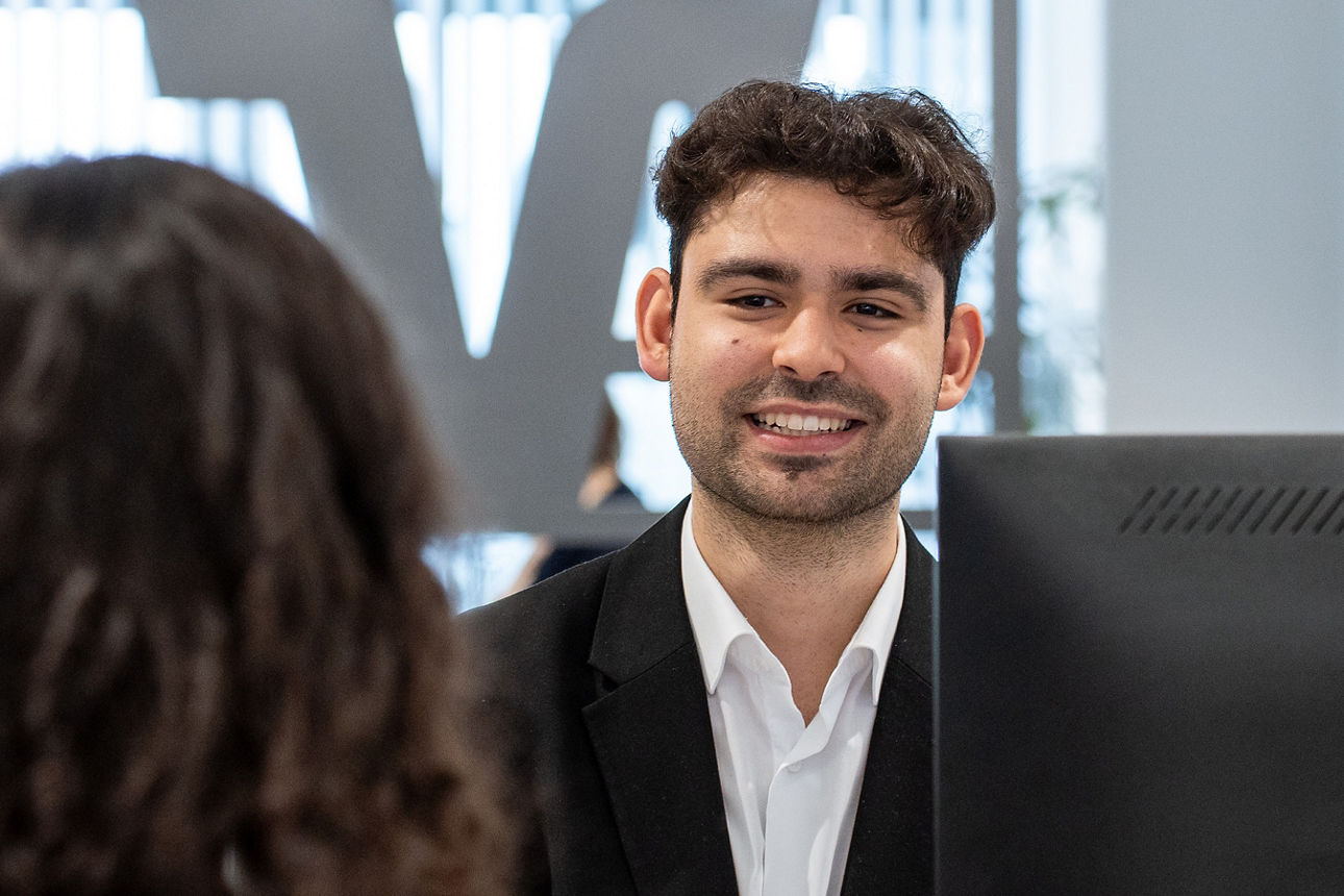 Auszubildender lächelt Kundin am Schalter an - im Hintergrund ist das VR-Logo zu sehen