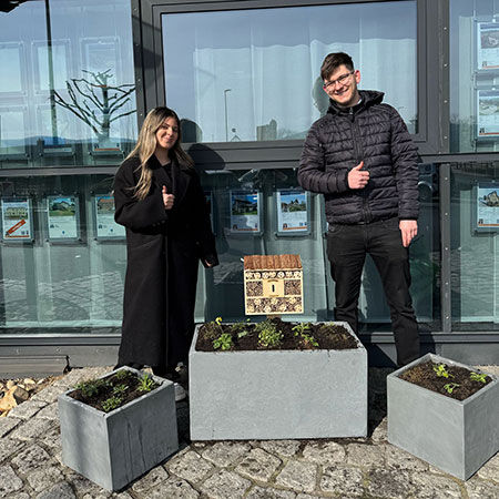 Zwei Auszubildende stehen vor einem Bankgebäude und präsentieren ihre gepflanzten Blumenkübel.