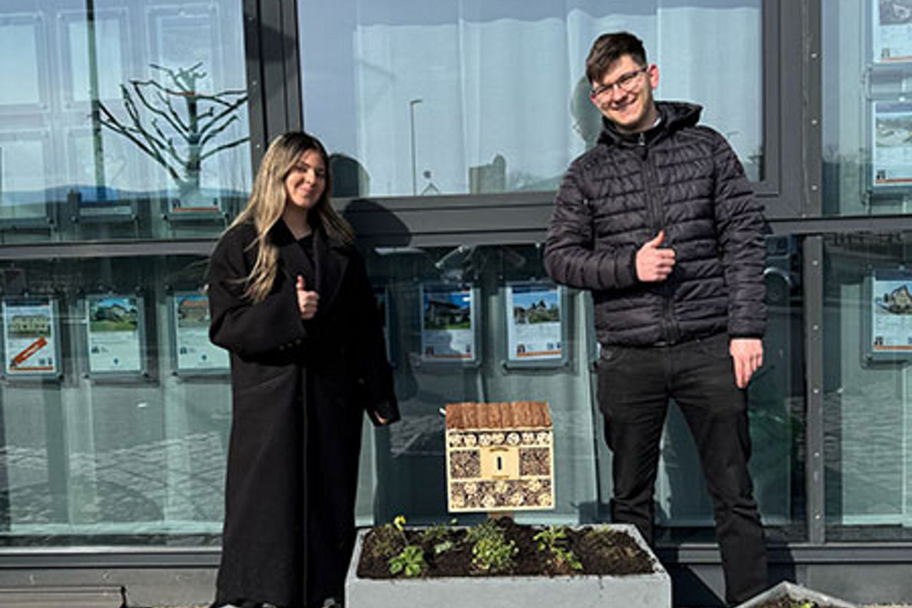 Zwei Auszubildende stehen vor einem Bankgebäude und präsentieren ihre gepflanzten Blumenkübel.