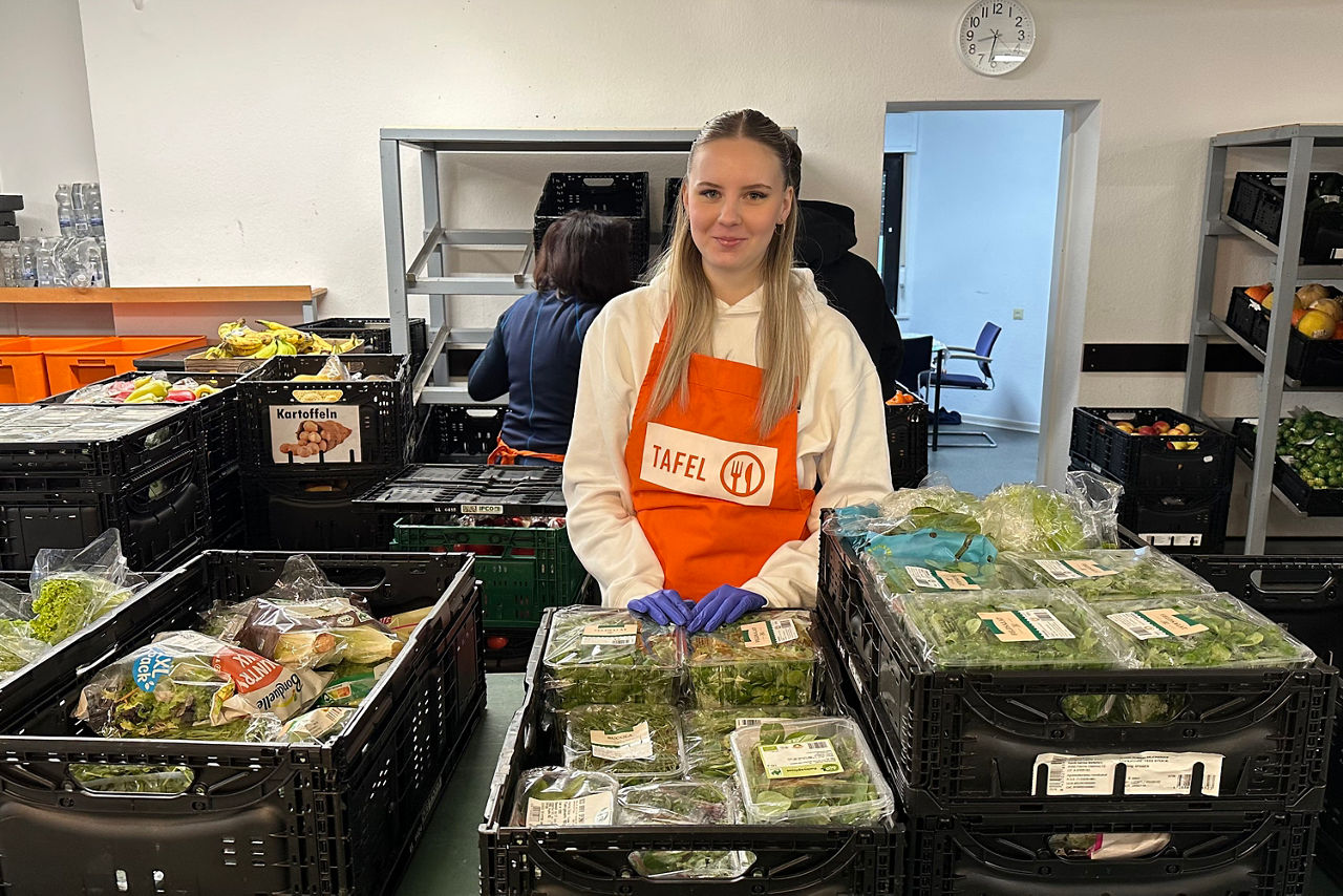 Frau mit oranger Schürze und blauen Handschuhen steht hinter Kisten mit verpacktem Gemüse in einem Lagerraum
