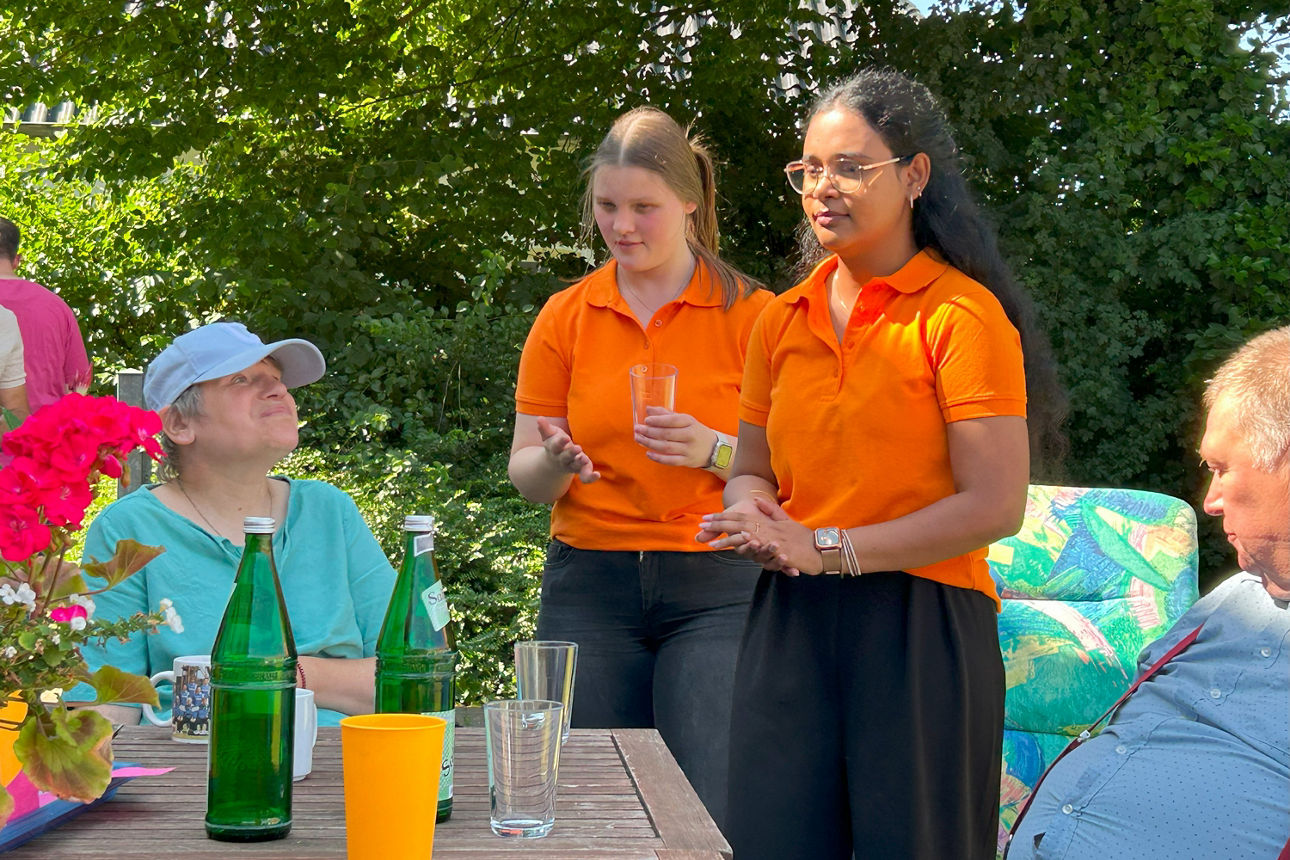 Zwei Frauen stehen vor einem Tisch und unterhalten sich mit zwei weiteren Personen am Tisch.
