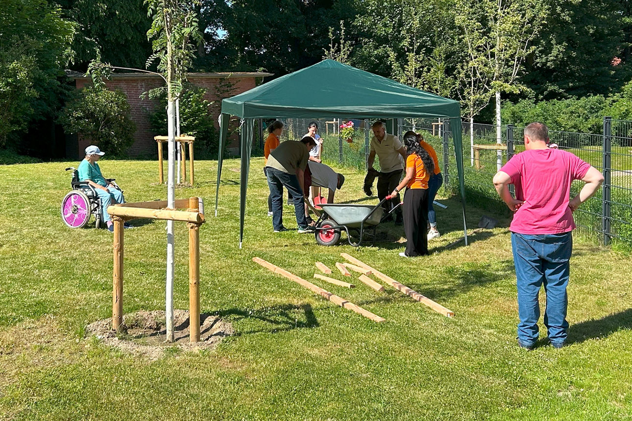 Eine Gruppe Menschen steht unter einem Pavillon und befüllt den Barfußpfad mit Erde aus einer Schubkarre. 