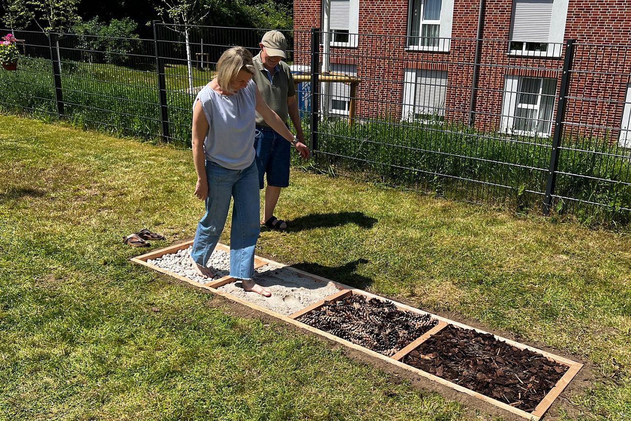 Eine Frau benutzt einen Barfußpfad (Stein, Sand, Tannenzapfen, Rindenmulch) und ein Mann steht seitlich hinter ihr.