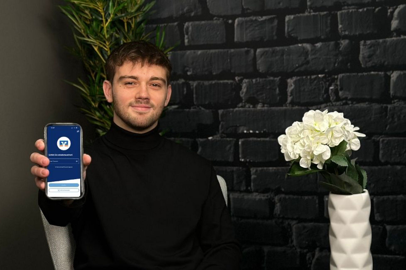 junger Mann sitzt lächelnd vor der Kamera mit Smartphone in der Hand auf welchem die VR Banking App geöffnet ist