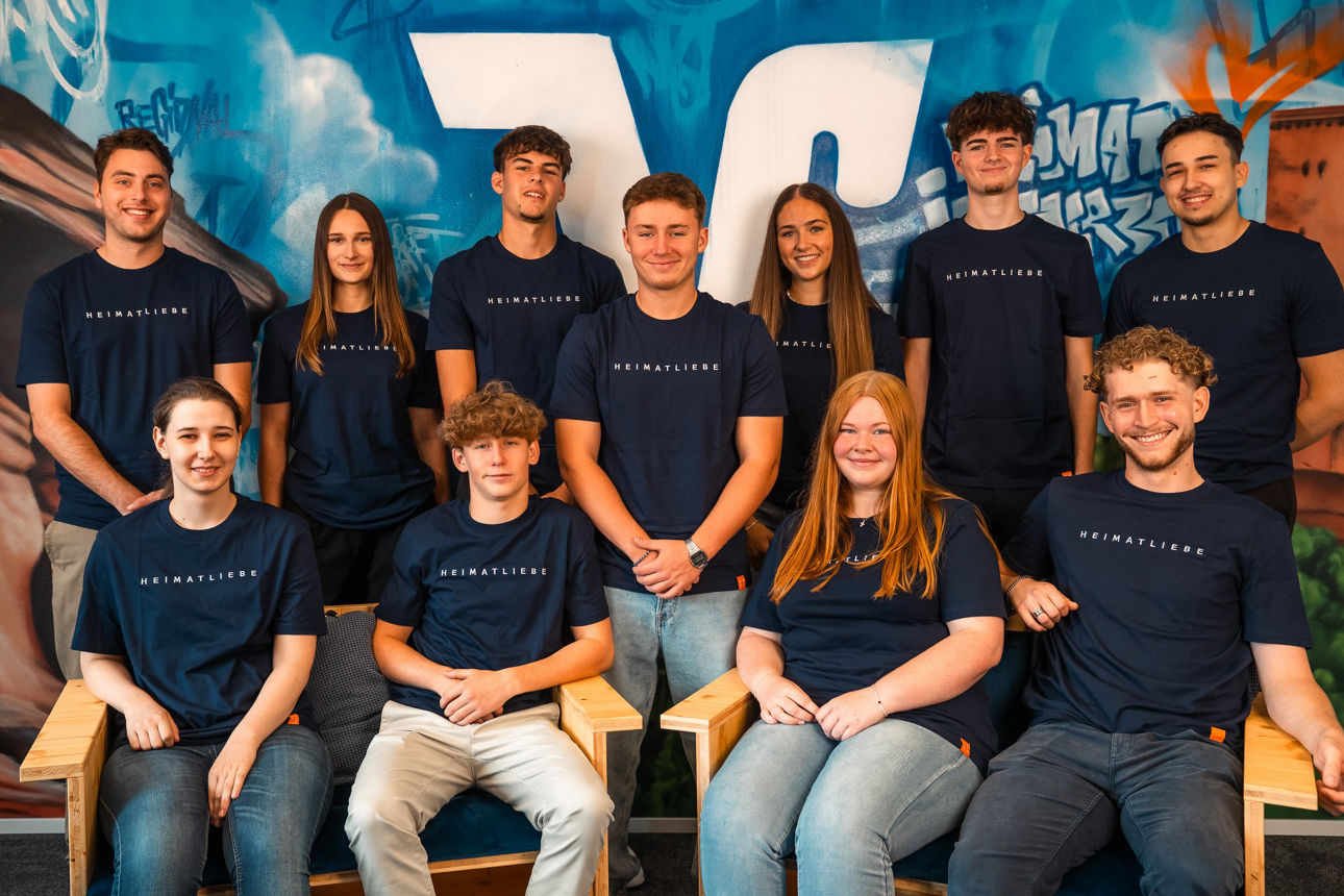 Zehn Personen in blauen T-Shirts vor einer bunt gestalteten Wand, sitzend und stehend