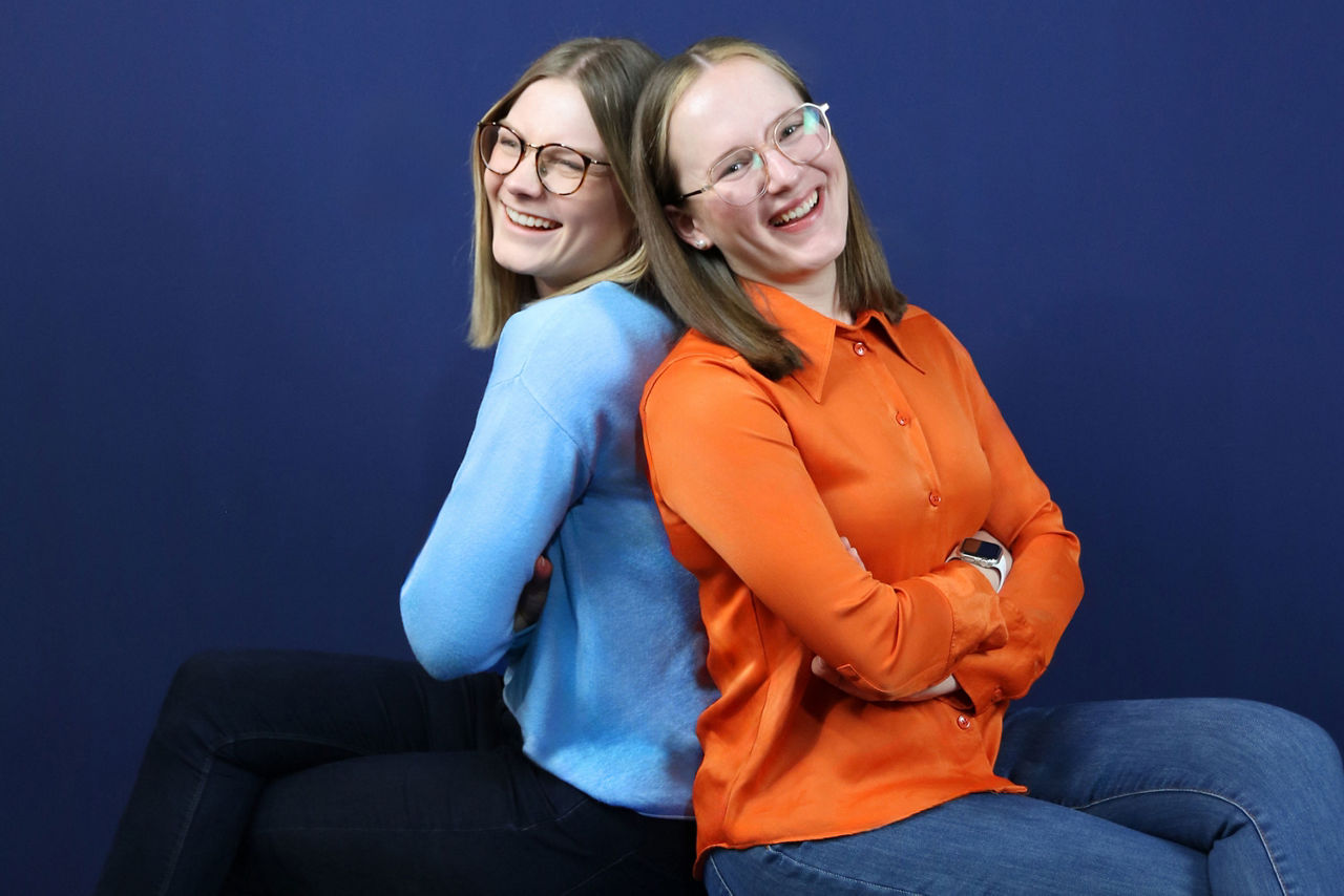Zwei Auszubildende der VR Bank Main-Kinzig-Büdingen sitzen Rücken an Rücken vor einem blauen Hintergrund und lachen herzlich. 
