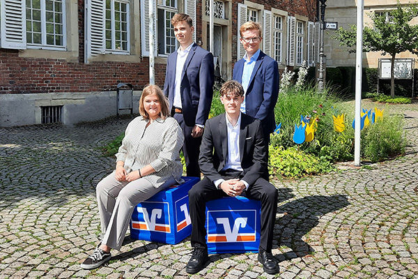 Vier junge Erwachsene stehen und sitzen vor einem Backsteingebäude.