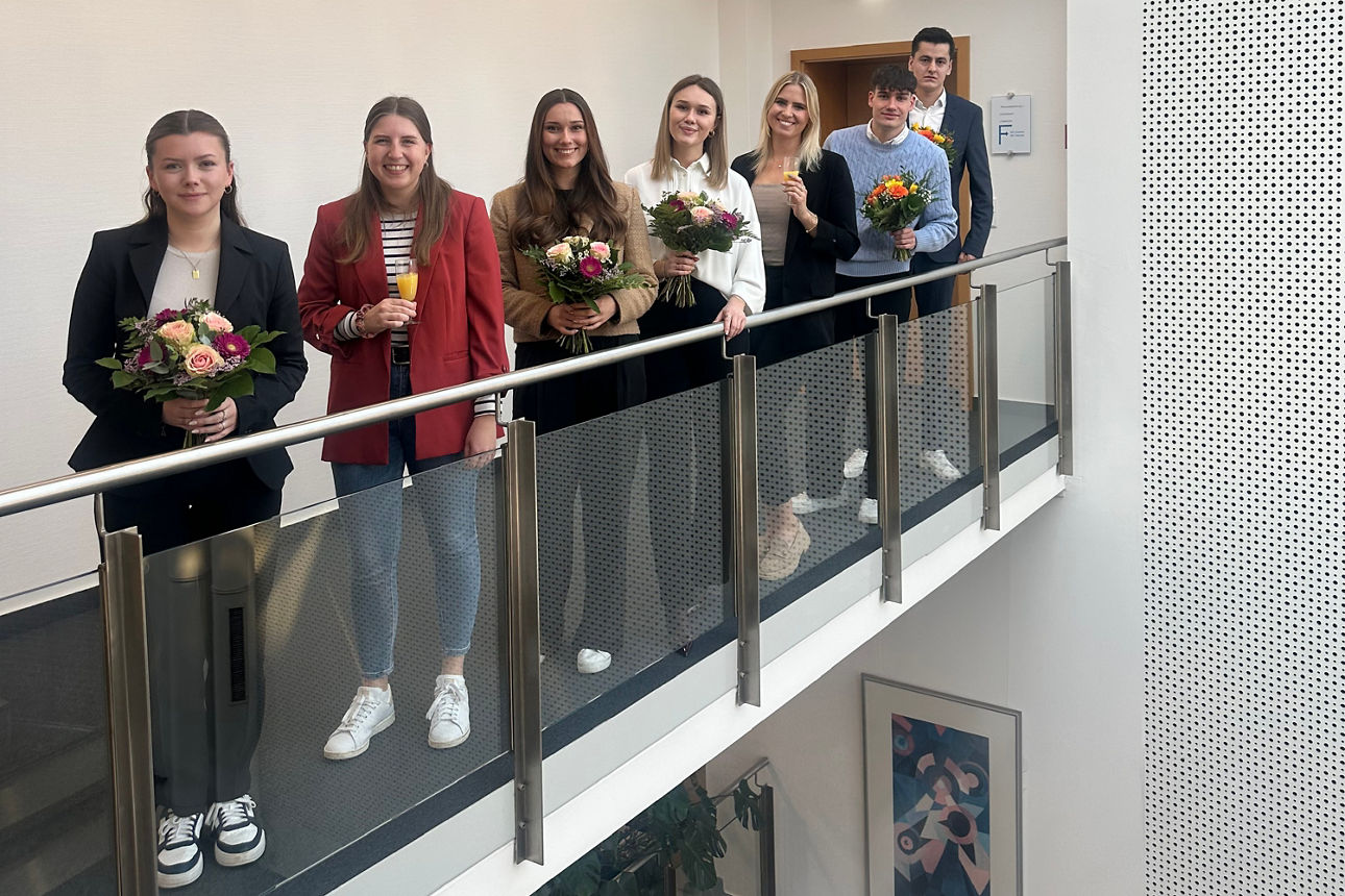 Sieben Personen stehen nebeneinander an einem Geländer in einem Innenraum, einige halten Blumensträuße