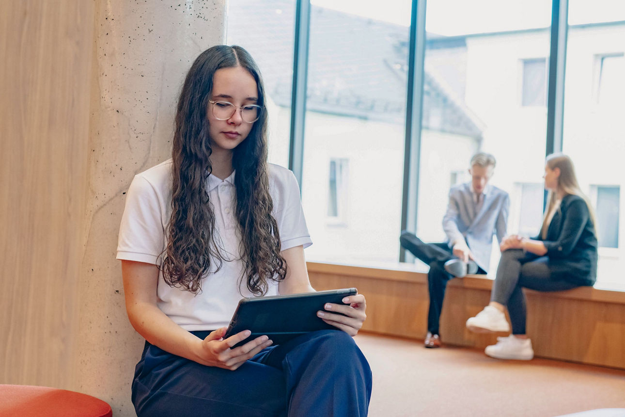 Newsletter - Abbildung einer jungen Frau mit dunklen langen Haaren und Tablet in der Hand, im Hintergrund sitzen 2 Personen