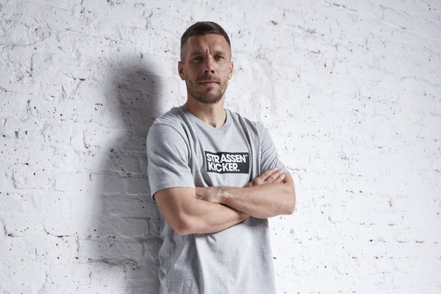 Testimonial Lukas Podolski steht vor einer hellgrauen Wand mit verschränkten Armen und lächelt in die Kamera