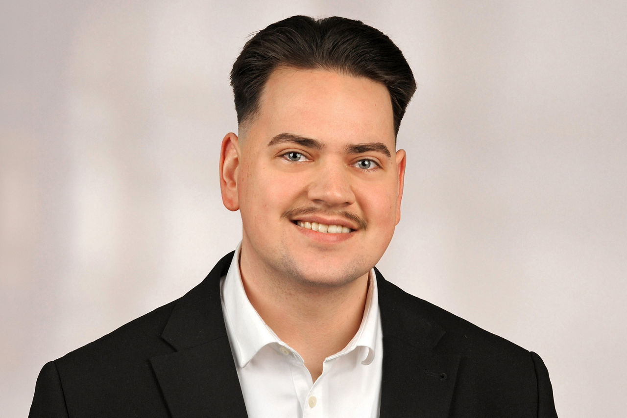 Benjamin Böhm hat braue kurze Haare und einen Oberlippenbart. Er trägt einen Anzug