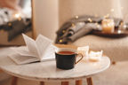 Tasse Tee mit aufgeschlagenem Buch und brennenden Duftkerzen auf einem Marmortisch vor einem Sessel und warm leuchten