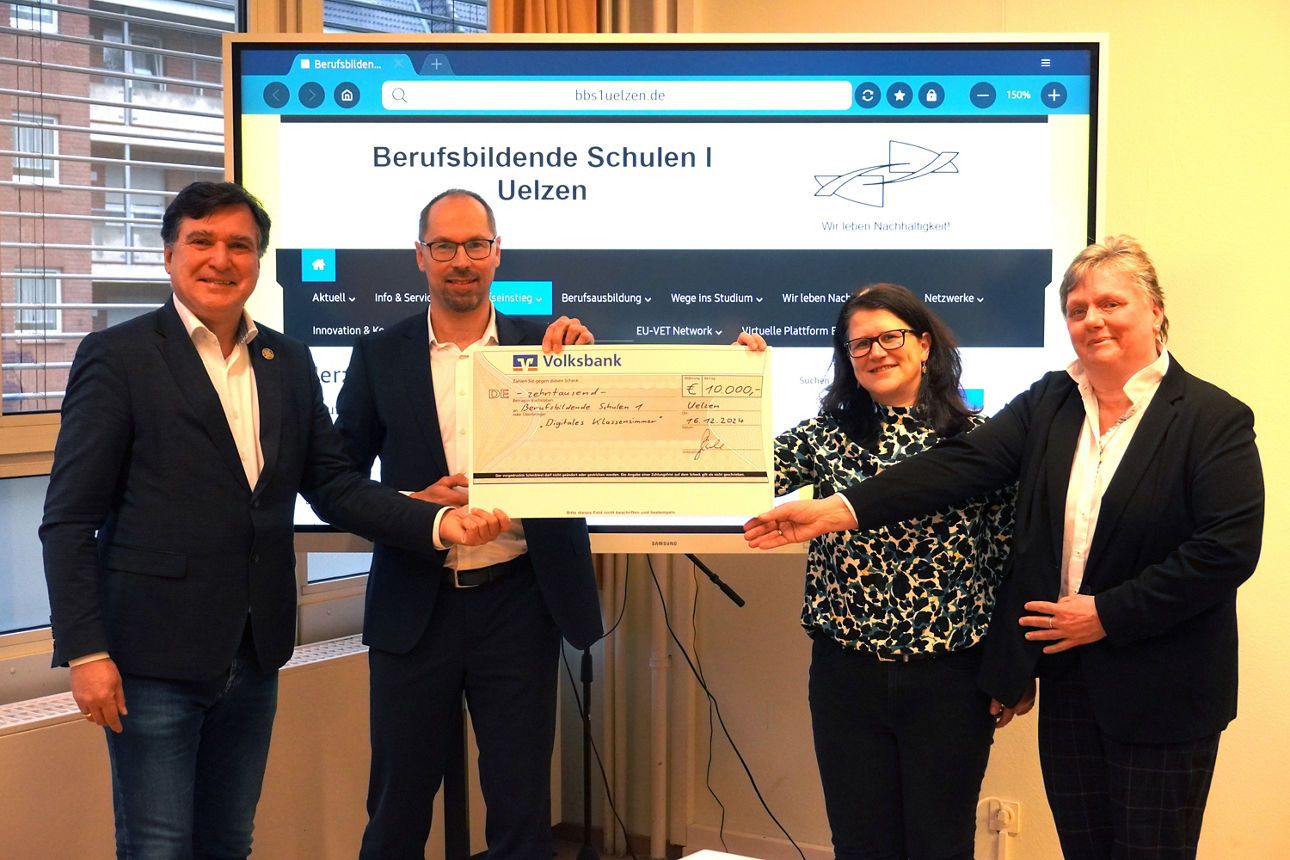 Zwei Männer und zwei Frauen stehen mit einem Spendencheck vor einer digitalen Tafel