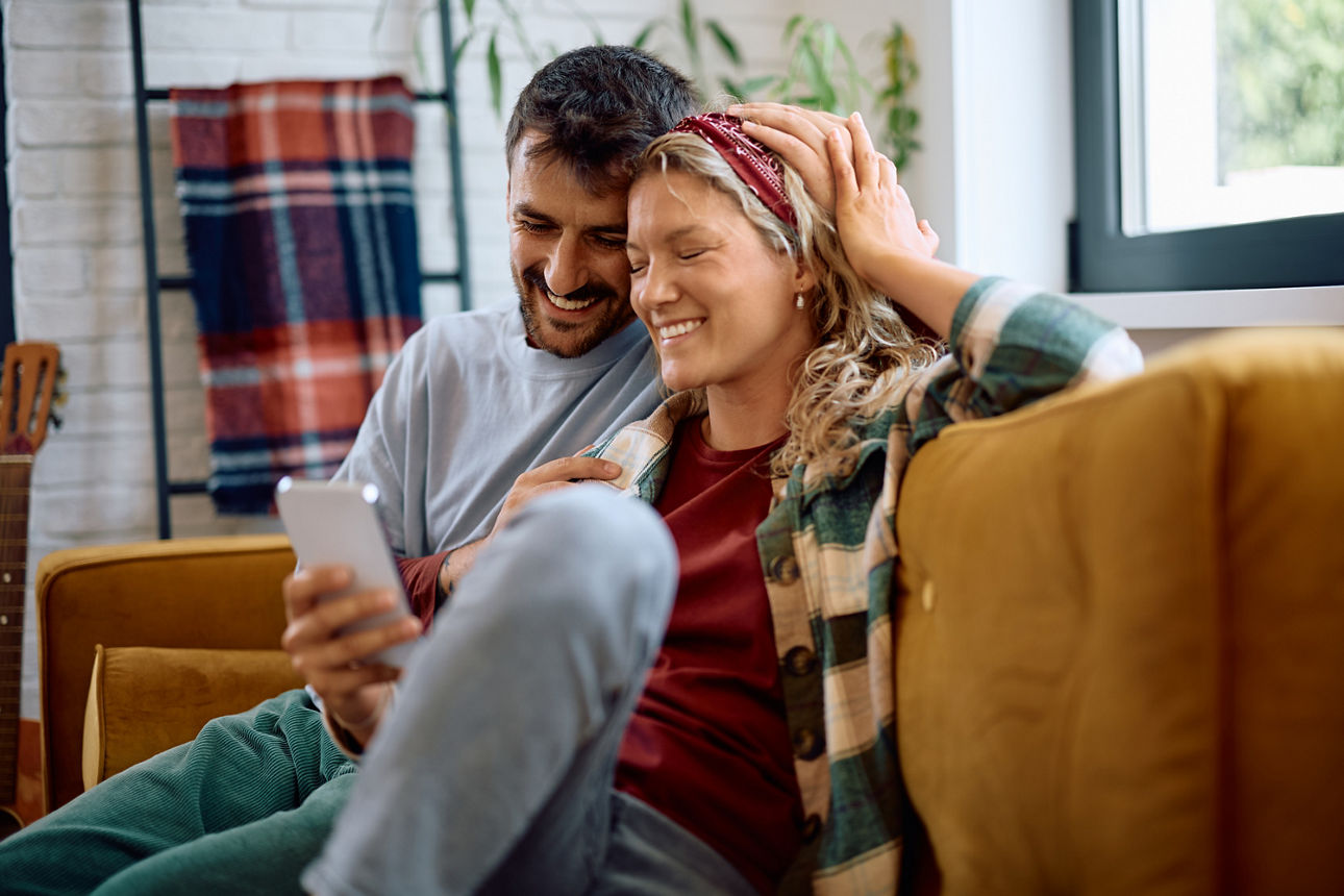 Fröhliches Paar sitzt gemeinsam auf dem Sofa und schaut auf ein Handy.