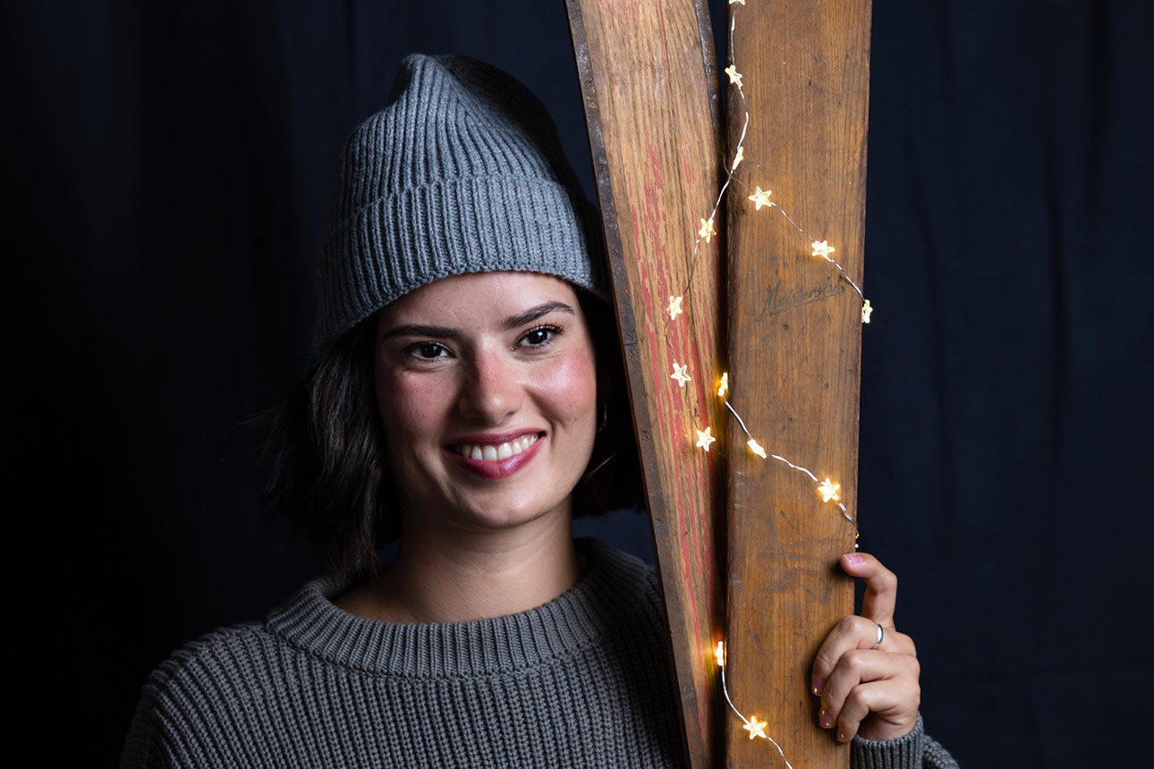 Junge Frau mit Mütze und Stickpulli hält zwei alte Holz-Ski mit einer Lichterkette und lächelt in die Kamera. 