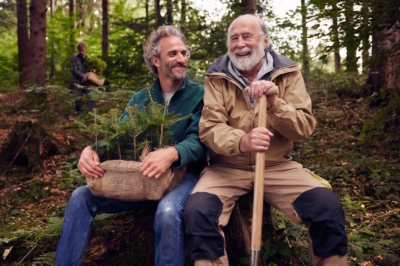 Zwei lachende Männer im Wald beim Bäume pflanzen mit Spaten und Setzlingen