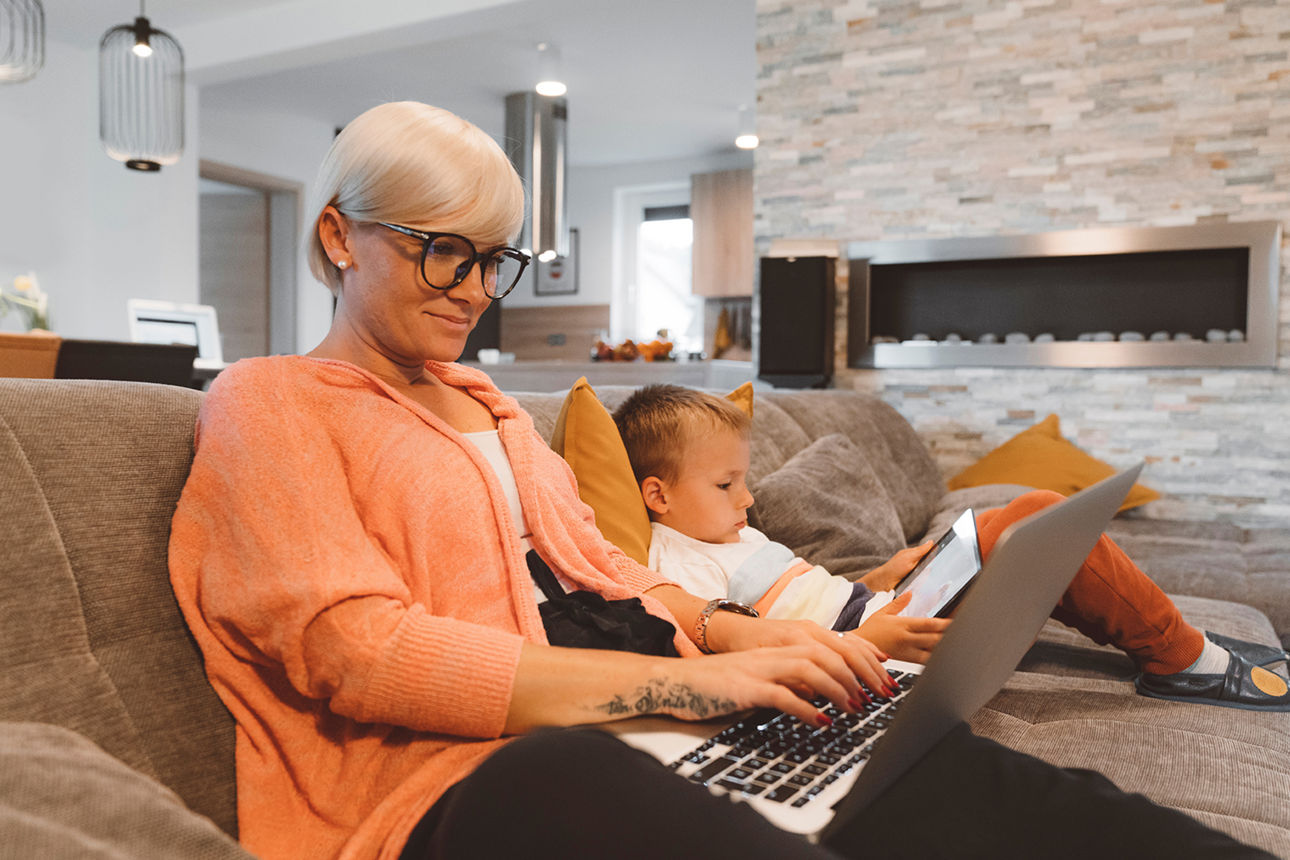 Termin Privatkunden - Seniorin sitzt auf dem Sofa, hat Laptop vor sich und schaut begeistert aufs Handy