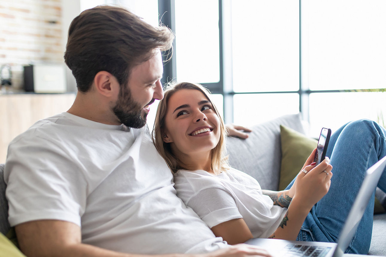Junges Pärchen sitzt auf dem Sofa und lächelt sich gegenseitig an, sie mit Handy in der Hand, er mit einem Laptop