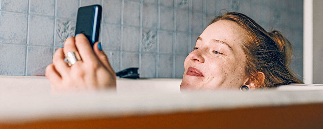 Junge Frau liegt in der Badewann und schaut lächeldn auf ihr Smartphone