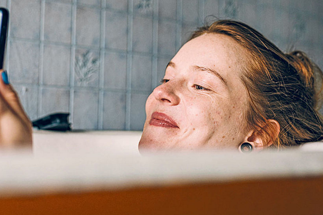 Junge Frau liegt in der Badewann und schaut lächeldn auf ihr Smartphone
