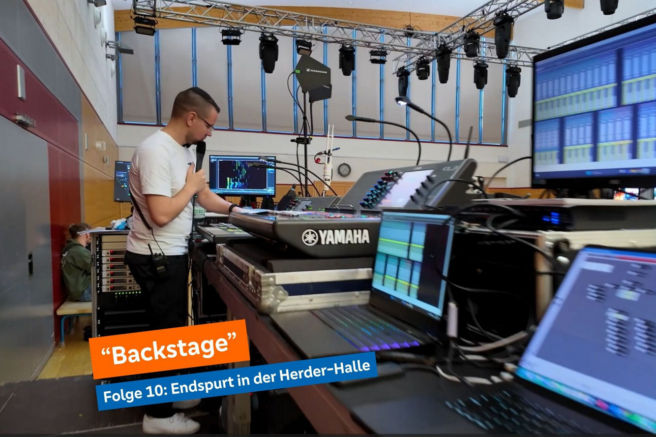 Tontechnik-Pult mit vielen Bedienelementen und Bildschirmen, Mann im T-Shirt bedient einen Schalter und spricht ins Mikro