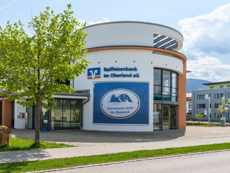 Die moderne Fassade der Raiffeisenbank im Oberland eG in Bad Tölz mit einem großen blauen Logo, umgeben von Bäumen und einer Straße.