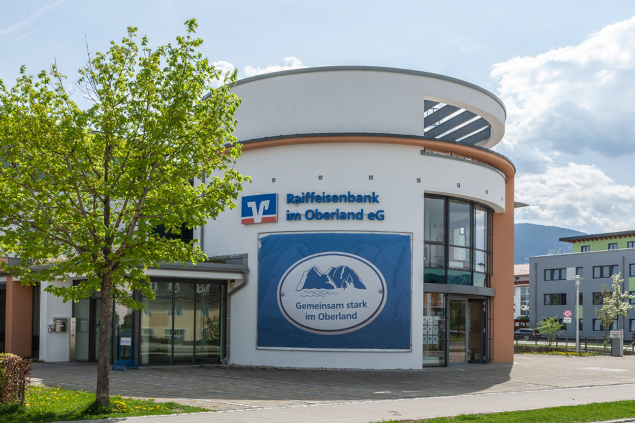 Ein hoher, moderner Bau der Raiffeisenbank im Oberland eG in Bad Tölz, mit einem großen Bergpanorama-Logo an der Fassade, umgeben von Bäumen und einer Straße.