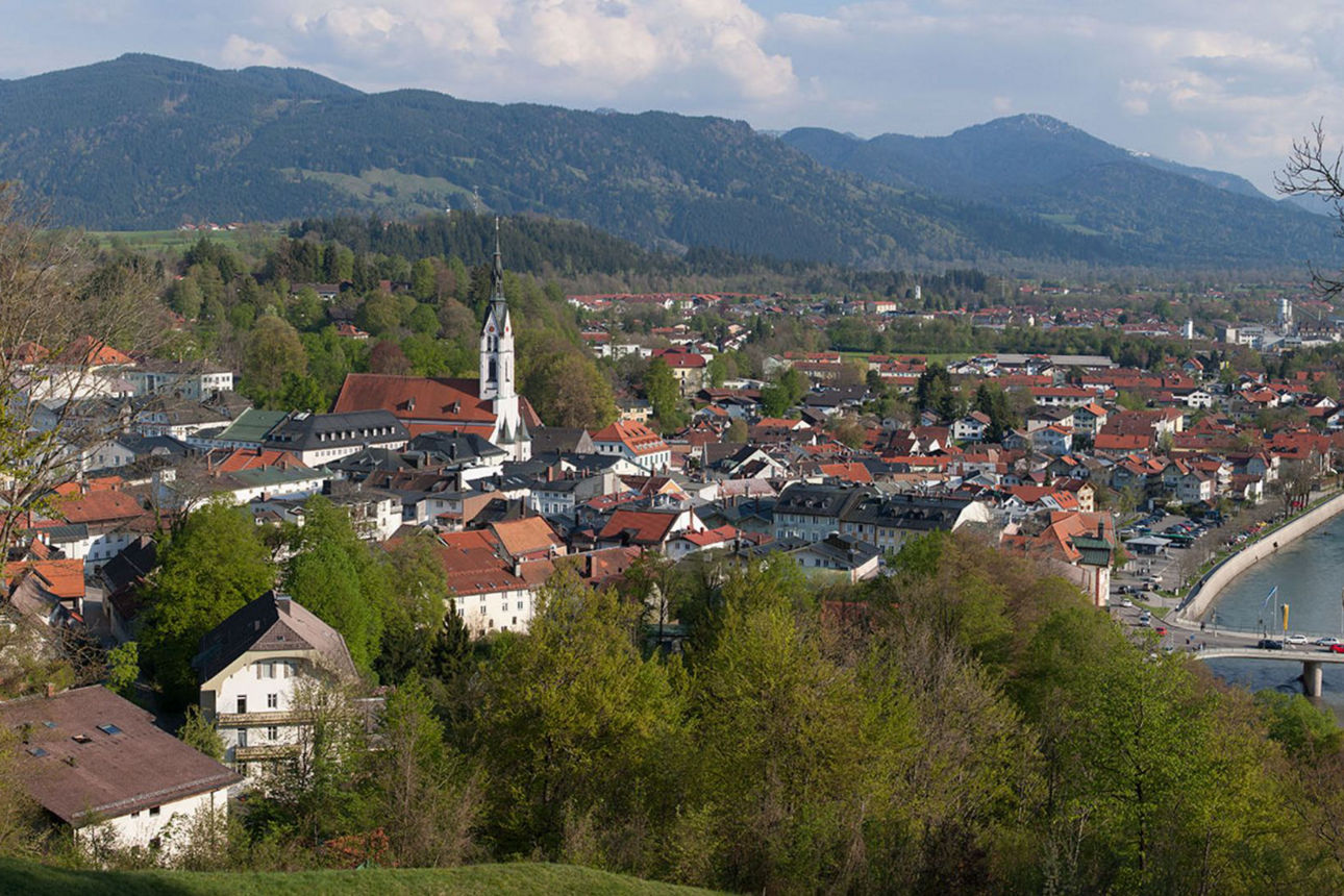 Eine erhöhte Ansicht der Stadt Bad Tölz, die sich entlang eines Flusses und einer Brücke erstreckt. Zahlreiche Häuser mit roten Dächern sind zu sehen, sowie eine große Kirche mit einem weißen Turm. Im Hintergrund erheben sich bewaldete Berge unter einem teilweise bewölkten Himmel.