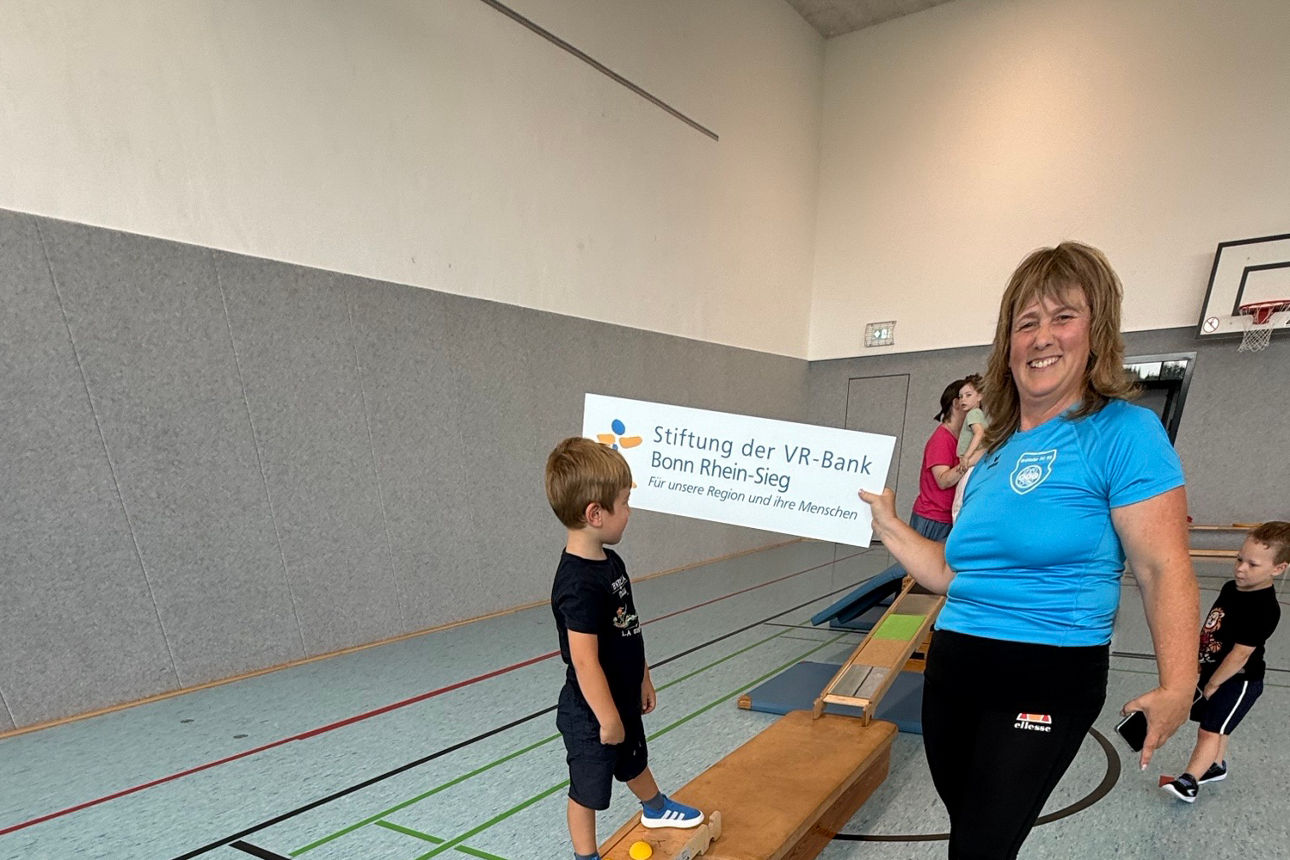 Kind balanciert in der Turnhalle auf einem Holz-Balancebalken, daneben steht eine Trainerin und zeigt auf ein Schild.