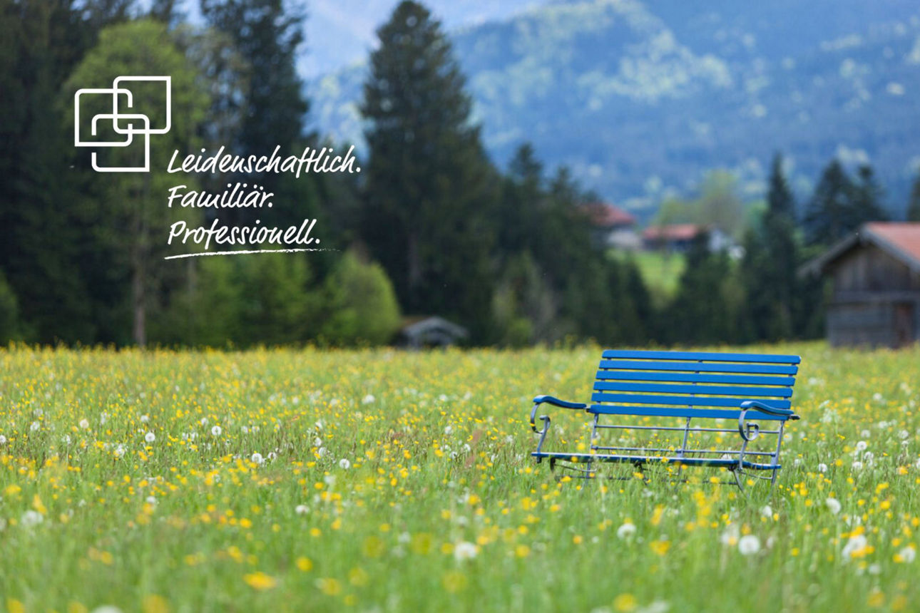 Eine blaue Holzbank steht mitten auf einer grünen Wiese mit gelben und weißen Blumen. Im Hintergrund sind Berge und Bäume zu sehen. In der oberen linken Ecke ist ein weißes Logo mit dem Text "Leidenschaftlich. Familiär. Professionell." zu sehen.