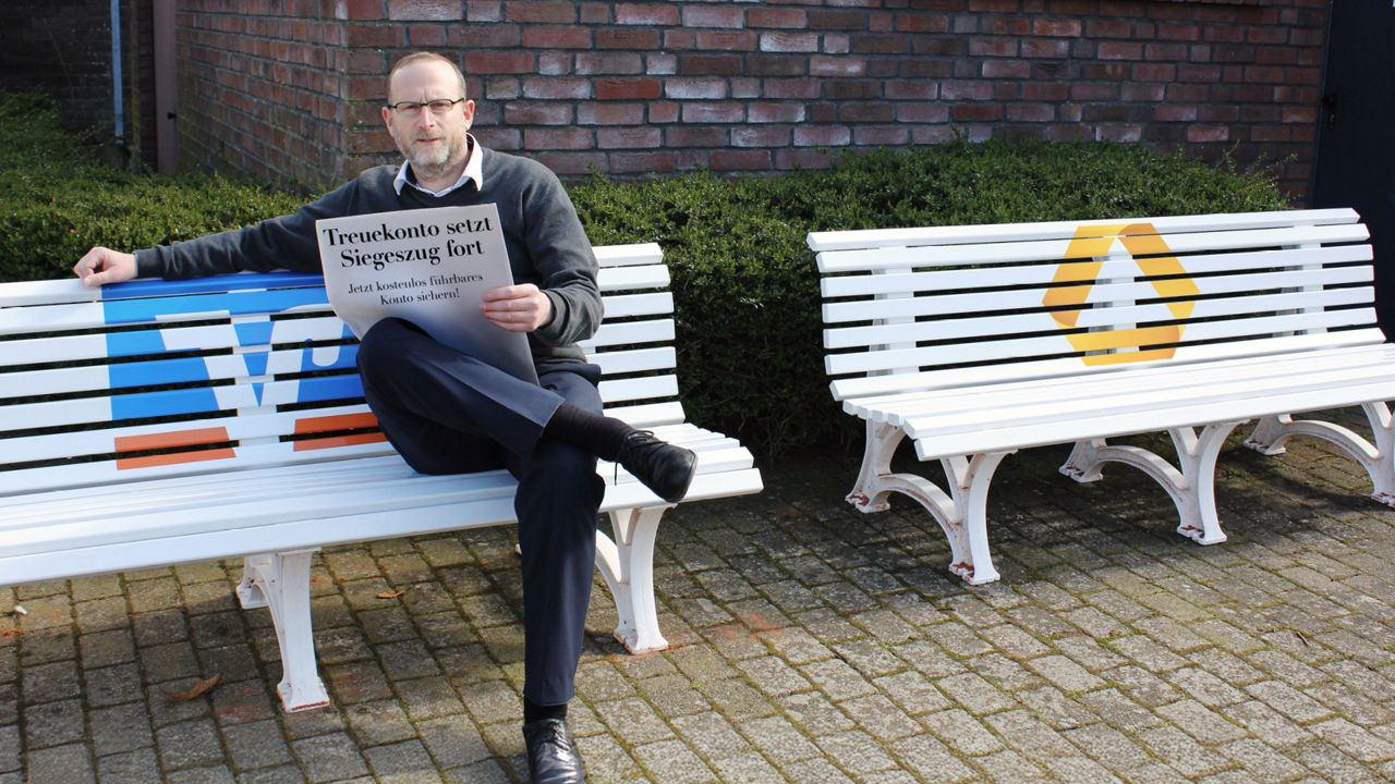 Mann sitzt mit Zeitung auf einer Bank mit Volksbank-Logo