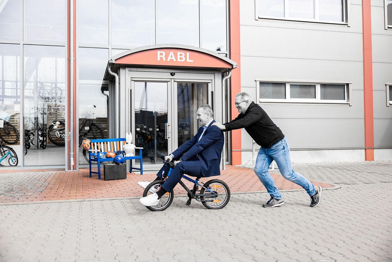 Ein Mann sitzt auf einem Kinderfahrrad und wird von einem anderen Mann geschoben.