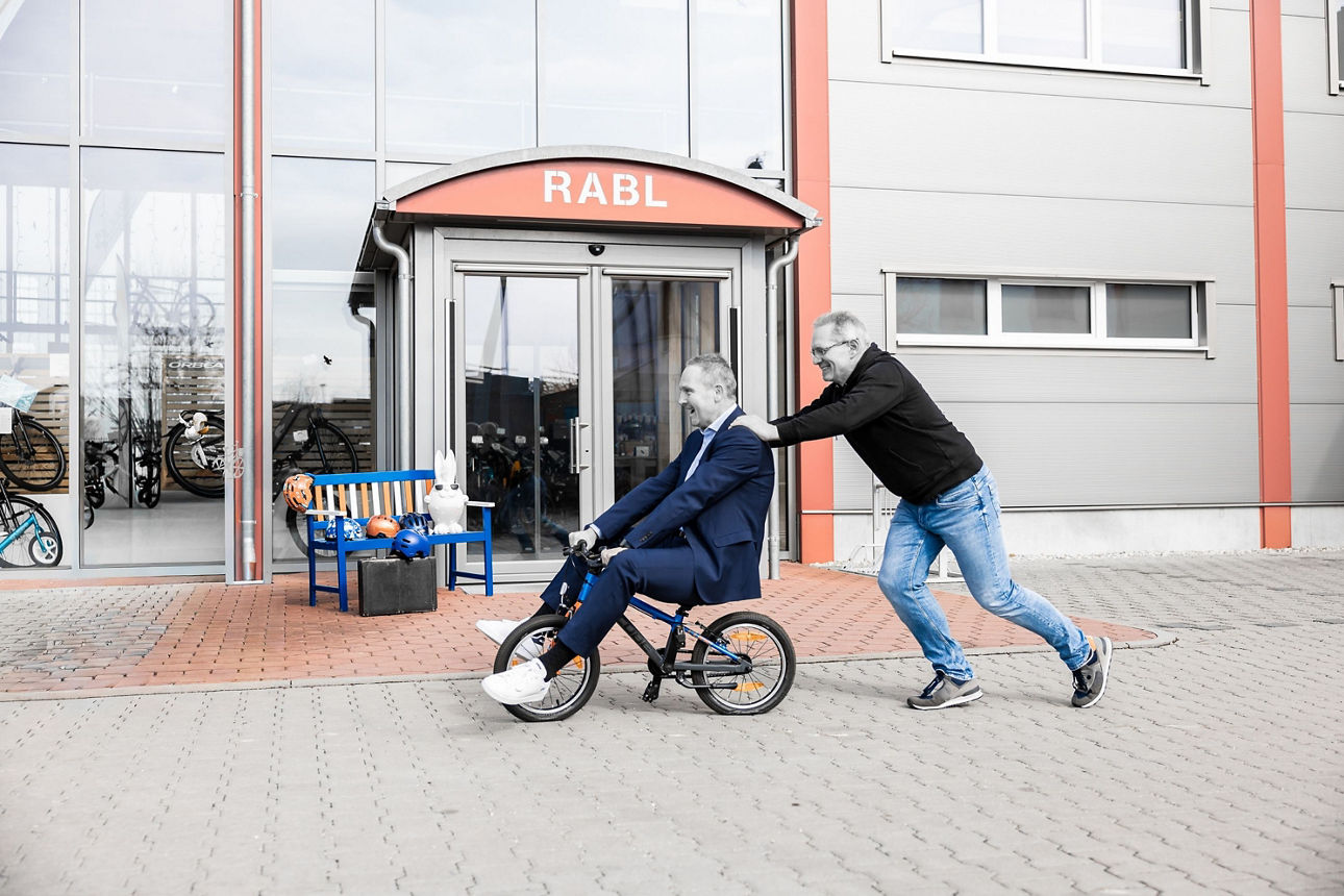 Ein Mann sitzt auf einem Kinderfahrrad und wird von einem anderen Mann geschoben.